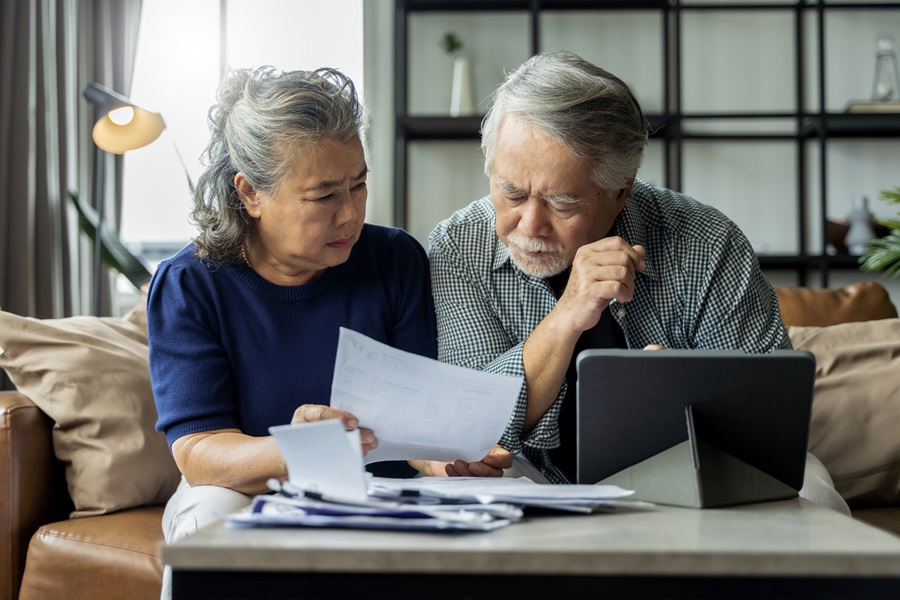 Older couple reviewing finances on paper and tablet computer in living room setting.